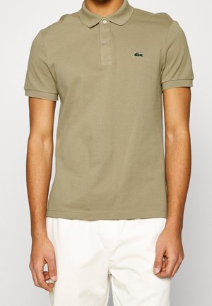 Mann trägt khakifarbenes Polo-Shirt mit kurzen Ärmeln und kleinem Krokodil-Logo auf der Brust sowie weiße Hose vor einfarbigem Hintergrund.