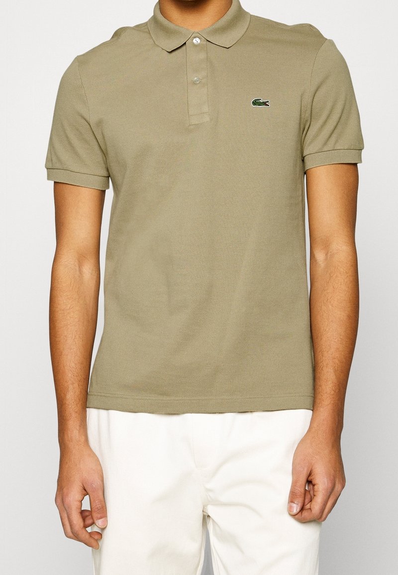 Homme portant un polo kaki à manches courtes avec un petit logo crocodile sur la poitrine et un pantalon blanc sur fond uni.