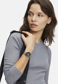 Ronde zwarte klok met zilveren metalen band, gedragen om een pols. Grijs geribbeld langemouwen shirt. Minimalistisch ontwerp, strakke lijnen en subtiele textuur.