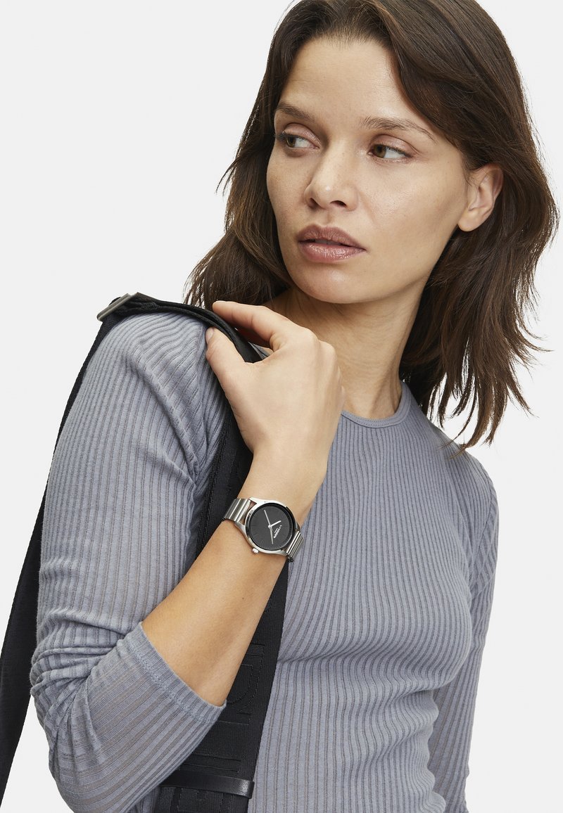 Ronde zwarte klok met zilveren metalen band, gedragen om een pols. Grijs geribbeld langemouwen shirt. Minimalistisch ontwerp, strakke lijnen en subtiele textuur.