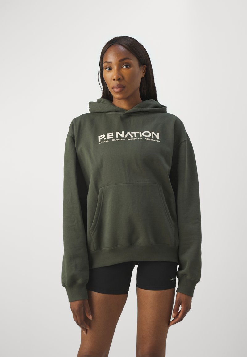 Felpa oversize in cotone verde con tasca frontale e logo stampato bianco. Caratterizzata da cappuccio con coulisse e polsini a coste. Indossata con pantaloni corti neri.