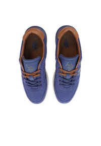 Lila sportskor med nätöverdel, bruna läderdetaljer, runda snören och en texturerad sula. Har en logotyp på tungan.