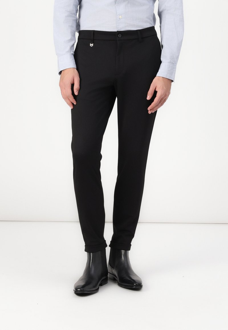 Pantalons slim noirs avec une texture lisse, dotés d'une fermeture à bouton et de poches latérales. Portés avec des bottines noires.