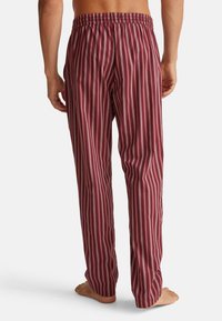 Man in maroon en witte verticaal gestreepte pyjamabroek, staande op blote voeten met de handen ontspannen naast het lichaam, gezien van achteren.