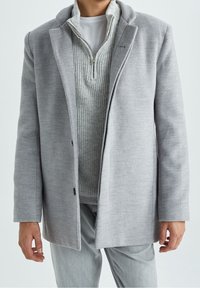 Abrigo de lana gris con un ajuste entallado, cierre con un solo botón y cuello con solapas, superpuesto sobre un suéter texturizado gris claro.