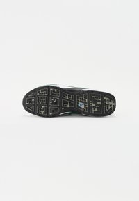 Semelle d'une chaussure de sport noire et verte avec un motif de bande de roulement et une petite étiquette sur la semelle intermédiaire, présentée sur un fond blanc uni.