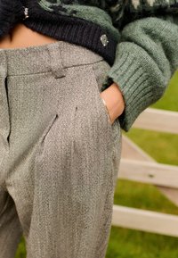 Šedé herringbone kalhoty s skladem, obsahující boční kapsy a vysoký pas, kombinované se zeleným pleteným svetrem.
