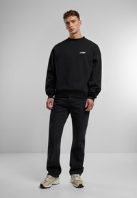 Czarny sweatshirt z okrągłym dekoltem, z prążkowanymi mankietami i dolnym wykończeniem, zestawiony z czarnymi dżinsami i beżowymi butami biegowymi. Minimalistyczny design z detalem logo.