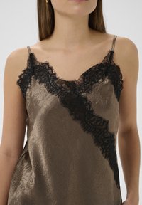 Brun satin cami med sort blonder langs halsudskæringen. Har tynde spaghettistropper og en tekstureret overflade.
