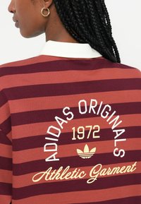 Prugasta atletska oblačila v temno rdeči in rjavi barvi; bela polo ovratnica; vsebuje besedilo "ADIDAS ORIGINALS 1972" in logo v krem in rumeni barvi.
