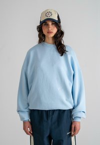 Helles blaues Sweatshirt mit Rundhalsausschnitt und gesenkten Schultern, kombiniert mit dunkelblauen Hosen mit weißen Seitenstreifen. Eine beige und schwarze Mütze mit Sternendesign.