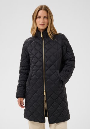 Femme portant un manteau matelassé noir jusqu'aux genoux avec une fermeture éclair dorée sur le devant et des poches latérales, debout devant un fond clair uni.