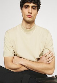 Beige Baumwoll-T-Shirt mit hohem Halsausschnitt und kurzen Ärmeln, mit einer glatten Textur und einer lockeren Passform. Model posiert mit verschränkten Armen.