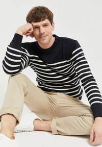 Maglione a righe blu navy e bianche con collo rotondo e dettagli di bottoni sulla spalla, abbinato a pantaloni beige arrotolati e sneakers bianche.