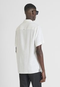 Camisa blanca de manga corta con un acabado texturizado, cuello abotonado y abertura en la parte trasera. Combinada con pantalones oscuros.