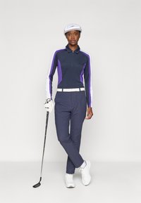 Marineblå golftrøye med lilla og hvite ermer, glidelåslukking; marineblå bukser; hvit belte og sko; holder en golfkølle. Flatt hvitt cap.