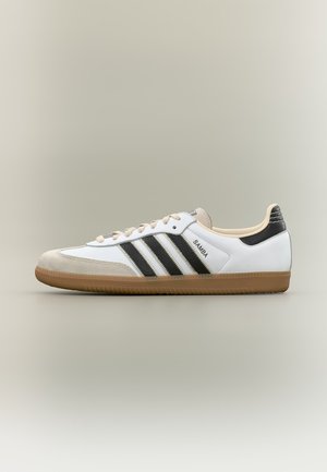 Biały sneaker Adidas Samba z czarnymi paskami, beżowym zamszowym noskiem, gumową podeszwą i czarnym detalem na pięcie na neutralnym tle.
