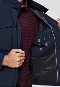 Marinblå vattentät jacka med flera inre fickor som har ikoner för förvaring. Mörk foder och kontrasterande vita detaljer längs dragkedjan.