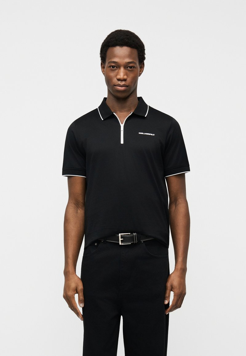 Νέος άνδρας που φοράει μαύρο κοντομάνικο πόλο με λευκό φερμουάρ και περιμετρικό σχέδιο, μαύρο παντελόνι και μαύρη ζώνη, στέκεται μπροστά σε μια απλή φόντο.