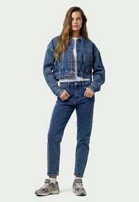 Džinsu jakas ar īsu dizainu un divām priekšējām kabatām, saskaņota ar augsto jostasvietu ziliem džinsiem. Modelis valkā pelēkas sporta apavas ar baltām akcentiem.