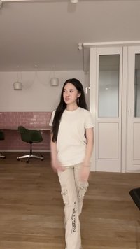 Femme aux longs cheveux noirs portant un t-shirt blanc et un pantalon cargo beige marchant à l'intérieur d'une pièce avec un mur carrelé rose et des chaises de bureau.