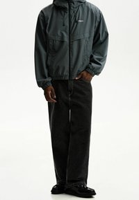 Man wearing dark green hooded zip jacket and loose black jeans with black shoes, standing with one hand in pocket on white background.