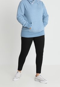 Persona con una sudadera de lunares azul claro, pantalones negros ajustados y zapatillas de plataforma blancas, de pie con las manos en los bolsillos.
