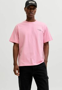 Pinkes Baumwoll-T-Shirt mit kurzen Ärmeln, Rundhalsausschnitt und einem kleinen Motiv auf der linken Brust. Kombiniert mit schwarzen Hosen und einer schwarzen Mütze.
