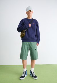 Navy sweatshirt with small red logo, green cargo shorts, and black/blue high-top sneakers. Gray cap and messenger bag complete the outfit.