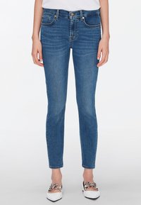 Jeans de denim azul ajustados, con cinco bolsillos, cierre delantero con botón y un ligero desvanecimiento. Combinados con zapatos blancos con acentos de cadena.