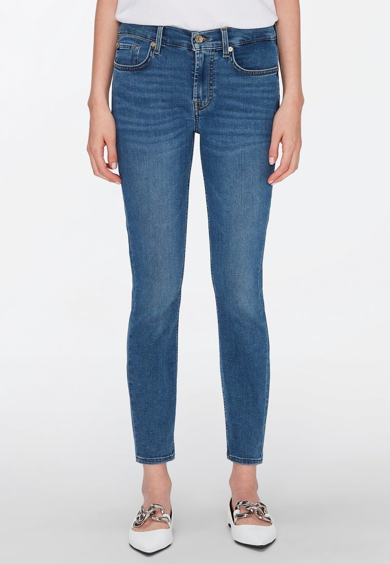 Jeans de denim azul ajustados, con cinco bolsillos, cierre delantero con botón y un ligero desvanecimiento. Combinados con zapatos blancos con acentos de cadena.