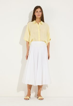 Femme portant un chemisier jaune clair boutonné, une jupe midi plissée blanche et des sandales ouvertes jaunes, debout devant un fond blanc.