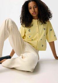 Geel gebreid cardigan met bloemenborduurwerk, korte mouwen en knoopsluiting, gecombineerd met roomkleurige relaxed-fit broek en zwarte schoenen.