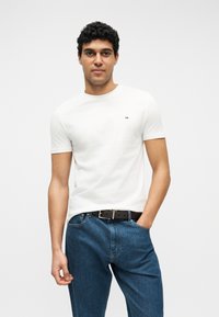 Νέος άνδρας με σγουρά μαλλιά, που φοράει λευκό κοντομάνικο μπλουζάκι και μπλε τζιν, στέκεται με το ένα χέρι στην τσέπη μπροστά σε απλό φόντο.