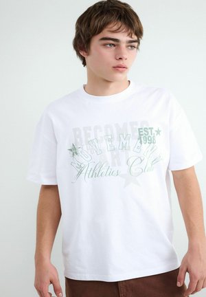 Young man with short brown hair wearing a white T-shirt with green and gray "Montemen Athletics Club" text and brown pants.