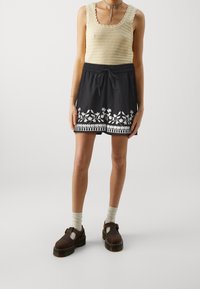 Pantaloncini neri con design floreale ricamato bianco e dettagli a righe, abbinati a un top leggero in crochet e scarpe marroni.