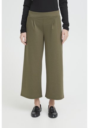 Mujer con pantalones cortos de pierna ancha de color verde oliva, camiseta negra de manga larga y mocasines negros, de pie frente a un fondo liso.
