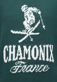 Zelena pletena jopica z belim dizajnom smučarskega lika in besedilom "CHAMONIX Francija" v strakih in kurzivnem tisku.