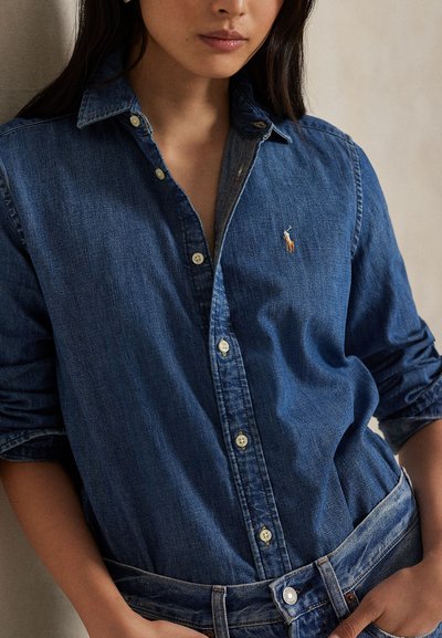 Femeie îmbrăcată cu o cămașă din denim albastru cu nasturi, cu mânecile suflecate și un mic logo brodat pe piept, asortată cu pantaloni din denim de aceeași culoare.