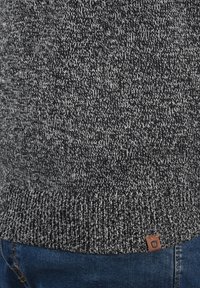 Pull en tricot dans des nuances de gris et de noir, avec un motif texturé et une étiquette en cuir marron au bas. Manches et bord inférieur côtelés et doux.