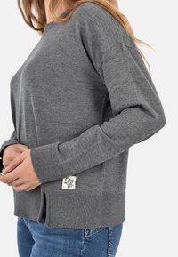 Maglione grigio lavorato a maglia con maniche lunghe, polsini a coste, spacchi laterali e una piccola patch in tessuto con logo nella parte inferiore sinistra.