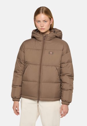 Veste rembourrée marron avec capuche, design matelassé, fermeture éclair à l'avant et poignets élastiques. Le modèle se tient devant un fond blanc uni.