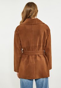 Giacca in suede marrone con vita regolabile, colletto ampio e dettagli di cucitura visibili. Il materiale appare morbido con una texture liscia.