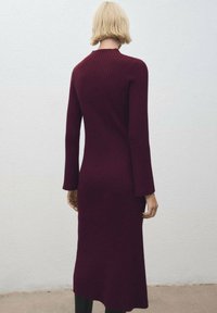 Abito in maglia a costine bordeaux con maniche lunghe e silhouette aderente, caratterizzato da un collo alto e lunghezza a metà polpaccio.