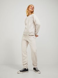 Felpa grigia chiara e pantaloni della tuta abbinati, entrambi realizzati in tessuto morbido, con polsini a coste e un piccolo dettaglio del logo. Sneakers nere completano il look.