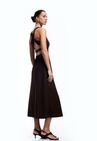 Vestido maxi marrón sin mangas con un corsé ajustado, espalda al aire y detalles de tirantes ajustables. Combinado con zapatos de tacón negro con tiras.