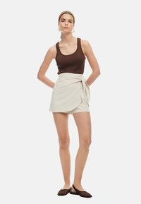 Bruine geribde tanktop, beige wikkelshorts met strikdetail en bruin gevlochten instapschoenen. Model staat rechtop tegen een eenvoudige witte achtergrond.
