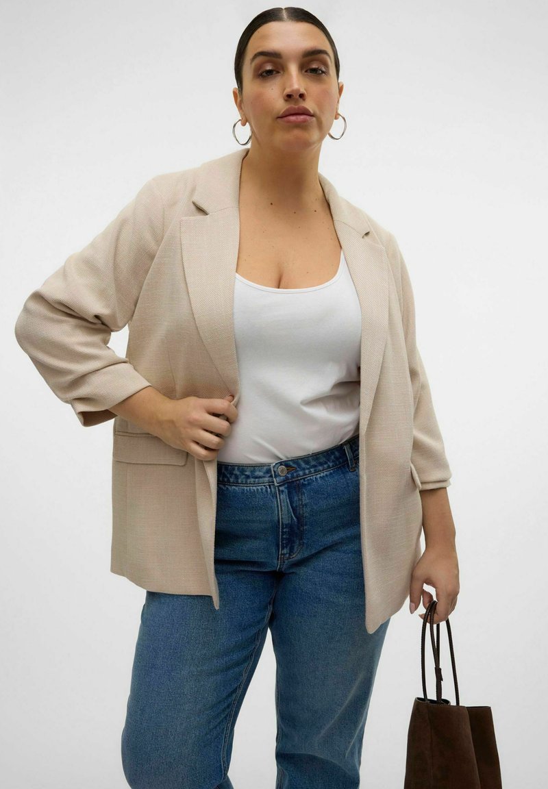 Femme portant un blazer beige, un débardeur blanc, un jean bleu, tenant un sac à main marron avec des boucles d'oreilles en anneau sur un fond blanc uni.