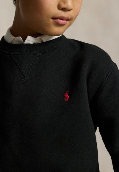 Persoană tânără purtând un pulover negru cu un mic logo de cal roșu brodat pe piept peste o cămașă albă cu guler.