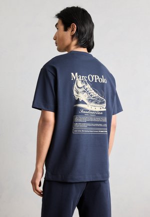 Homme portant un t-shirt bleu marine avec un grand graphisme vintage de chaussure de football et le texte « Marc O’Polo » dans le dos.